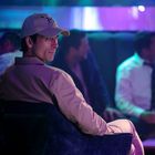 Mann mit Lächeln und Baseballkappe in Lounge, gedämpftes Licht, Menschen im Hintergrund in Gespräch vertieft.