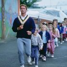 Ein Mann in Sportkleidung führt eine Gruppe von Kindern. Sie gehen auf einer Straße. Links ist eine bunte Wand zu sehen.