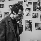 Eine Person mit Brille raucht, in einem Raum voller Schwarz-Weiß-Fotos an einer Wand.