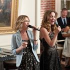 Zwei Frauen singen mit Mikrofonen bei einer Feier. Im Hintergrund Gemälde und Gäste.