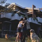 Eine Familie umarmt sich im Garten vor einem modernen Haus. Ein Hund sitzt daneben, ein anderer läuft im Hintergrund.