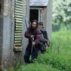 Zwei Personen schleichen mit Bögen entlang einer alten, überwucherten Mauer. Grauer Himmel, grüne Vegetation umgibt sie.