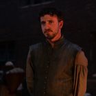 Ein Mann in historischer Kleidung steht nachdenklich in einem dunklen, mittelalterlichen Hof mit gedämpfter Beleuchtung.