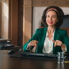 Frau tippt lächelnd an einem Schreibtisch mit Computer, Wasser und Büroartikeln, trägt grünen Blazer und Haarband.