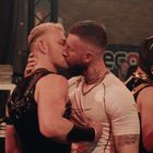 Zwei Männer küssen sich innig, daneben steht eine Person in Hundemaske. Im Hintergrund sind Regale und ein Plakat zu sehen.