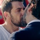 Zwei Männer küssen sich innig. Einer trägt ein weißes T-Shirt, der andere einen Anzug. Hintergrund: rotes Fahrzeug.