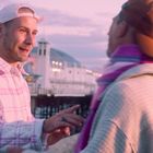 Text: "BRIGHTON PALACE". Zwei Personen unterhalten sich bei Sonnenuntergang am Pier. Links trägt jemand rosa Karohemd.