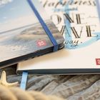 Text: "Happiness is just ONE WAVE away." Zwei Notizbücher, eines mit Strandmotiv, das andere mit blauem Anker. Beige Stoffunterlage.