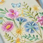 Eine Stickerei von bunten Blumen und einem Schmetterling auf hellem Stoff, detailliert und farbenfroh gestaltet.