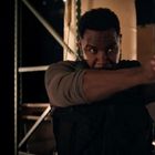 Ein Mann in einem Lager, der eine Pistole abfeuert. Kartons stapeln sich im Hintergrund.