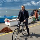 Ein Mann schiebt ein Fahrrad am Hafen, im Hintergrund Boote und Fischer vor einer stürmischen Meereskulisse.
