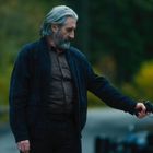 Ein älterer Mann mit grauen Haaren hält draußen eine Waffe, neben einem geöffneten Autotür.