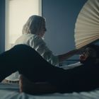 Zwei Personen auf einem Bett, eine reicht einem Getränk. Blaue Wände, zwei Bilder, hölzernes Dekorelement im Hintergrund.