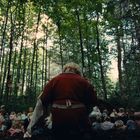 Ein Mensch in roter Kleidung kniet in einem Wald, umgeben von vielen Puppen, die auf dem Boden sind.