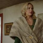 Eine Frau in einem Raum trägt eine elegante Fellstola, an der Wand sind dekorative Rahmen zu sehen.