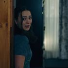 Eine Frau schaut ängstlich um eine Ecke in einem dunklen Raum mit Vorhängen und Holztüren.