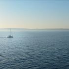 Ein Segelboot auf ruhigem Meer, im Hintergrund die Küstenlinie unter einem klaren, blauen Himmel.