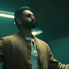 Ich kann keine Namen oder Texte erkennen. Ein Mann mit Bart und ausgebreiteten Armen steht in einer dunklen Umgebung.