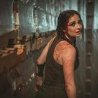 Eine Frau mit blutverschmiertem Gesicht und Kleidung schaut ängstlich zurück. Sie steht zwischen Paletten und Plastikfolien in einem engen Gang.