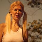 Ich kann die Texte nicht erkennen. Eine Frau mit blonden Zöpfen und einem weißen Top steht im Freien vor Bäumen.