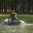 Ein Mann paddelt in einem Schlauchboot auf einem ruhigen Fluss, umgeben von Bäumen im Hintergrund.