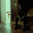 Keine Texte sichtbar. Eine Person sitzt nachdenklich auf einer hölzernen Treppe neben einem Fenster. Im Hintergrund ein heller Raum.