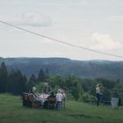 Einige Menschen sitzen an einem Tisch im Freien, umgeben von Hügeln und Wäldern, rechts steht ein geschlossener Sonnenschirm.