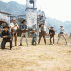 Sieben bewaffnete Cowboys in verschiedenen Posen vor einer alten Kirche, umgeben von Bergen.