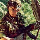 Ein Soldat in Tarnkleidung mit Gewehr, steht wachsam im Wald. Er trägt ein Stirnband und hat Tarnfarbe im Gesicht.