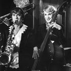 Drei Musiker: zwei spielen Saxophon und Kontrabass, die Dritte im Hintergrund Posaune. Alle in eleganter Abendkleidung.