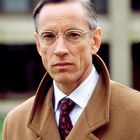 Ein Mann trägt einen braunen Mantel, Krawatte und Brille. Er hat kurzes Haar und blickt ernst. Der Hintergrund ist unscharf.