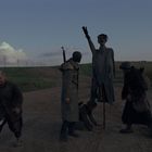 Kein Text vorhanden. Vier Personen auf einer unbefestigten Straße, eine hockt am Boden, eine Skulptur steht da.