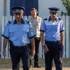 Zwei Polizisten in blauen Uniformen gehen ernst, während ein Mann im Hintergrund vor einem weißen Zaun steht.