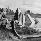 Zwei Personen sitzen in einem alten Holzboot am Ufer, im Hintergrund Wald und eine Kirche. Auf dem Boot steht "158".