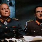 Zwei Männer in dunkellila Militäruniformen mit Abzeichen und Eisernem Kreuz sitzen an einem Tisch mit Papieren.