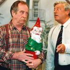 Zwei ältere Männer, einer mit kariertem Hemd hält einen Gartenzwerg, der andere gestikuliert. Hintergrund zeigt ein Haus.