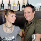 Ein Mann und eine Frau stehen lächelnd vor einer Bar mit Weinflaschen im Hintergrund.