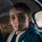 Ein Mädchen in einem Auto, ihr Gesicht ist im Fenster zu sehen. Reflektiert ist das Gesicht eines älteren Menschen.