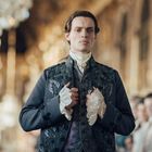 Eine Person in edlem, historisch anmutendem Gewand mit Spitzenmanschetten steht in einem prunkvollen Raum voller Kronleuchter.