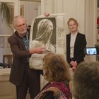 Ein Mann hält eine Zeichnung einer verschleierten Frau, umgeben von Menschen. Eine Kamera filmt die Szene.