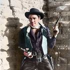 Ein Mann in Cowboy-Kleidung mit Hut hält zwei Revolver und steht vor einer Steinwand.