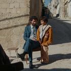 Ein Mann im Anzug sitzt auf einem Stuhl und spricht mit einem Jungen in brauner Jacke. Sie sind in einer sonnigen Gasse.