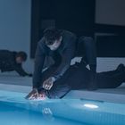 Eine dramatische Szene, in der ein Mann einen anderen am Rand eines Pools unter Wasser drückt, während eine weitere Person am Boden kauert.