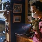 Ein Mensch mit Maske und Handschuh hält eine Taschenlampe. Im Raum sind Regale mit Geschirr, ein Ofen und Bilder an der Wand.