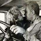 Zwei Personen in einem Fahrzeug, eng umschlungen, der Mann am Steuer, Nahaufnahme. Schwarz-Weiß-Fotografie.