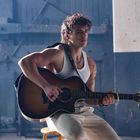 Ein Mann in einem weißen Tanktop sitzt und spielt Gitarre, im Hintergrund industrielle Kulisse.