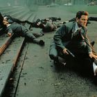 Ein Mann in Anzug kniet mit einer Pistole in der Hand über einem anderen Mann auf nassem Boden, umgeben von leblosen Körpern.