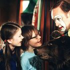 Ein Clown mit weiß geschminktem Gesicht ist von drei lachenden Kindern umgeben und hält einen Bärenkopf.