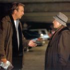 Zwei Personen stehen in einem Parkhaus, vertieft in ein Gespräch. Eine Person hält eine Plastiktüte mit Inhalt in der Hand.