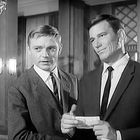 Zwei Männer in Anzügen stehen in einem eleganten Raum mit Holzverkleidung. Einer hält ein kleines Objekt in der Hand.